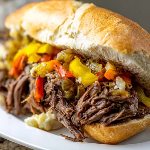 Tender slow-cooked Italian Beef Sandwich with giardiniera and roasted peppers, served alongside a small bowl of savory au jus for dipping.
