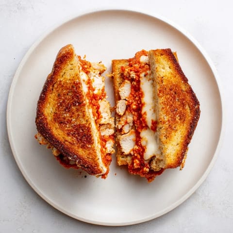 A close-up of a juicy Chicken Parm Grilled Cheese, cut diagonally to reveal the layered ingredients and vibrant basil garnish.  