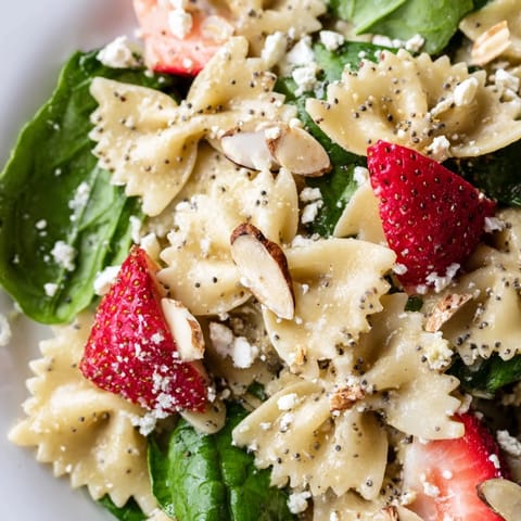 A close-up of Spring Spinach Strawberry Pasta Salad shows feta crumbles and basil over a vibrant bed of greens and fruit.